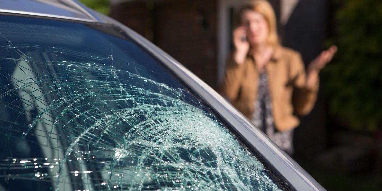 A woman on the phone while looking at a broken windshield.